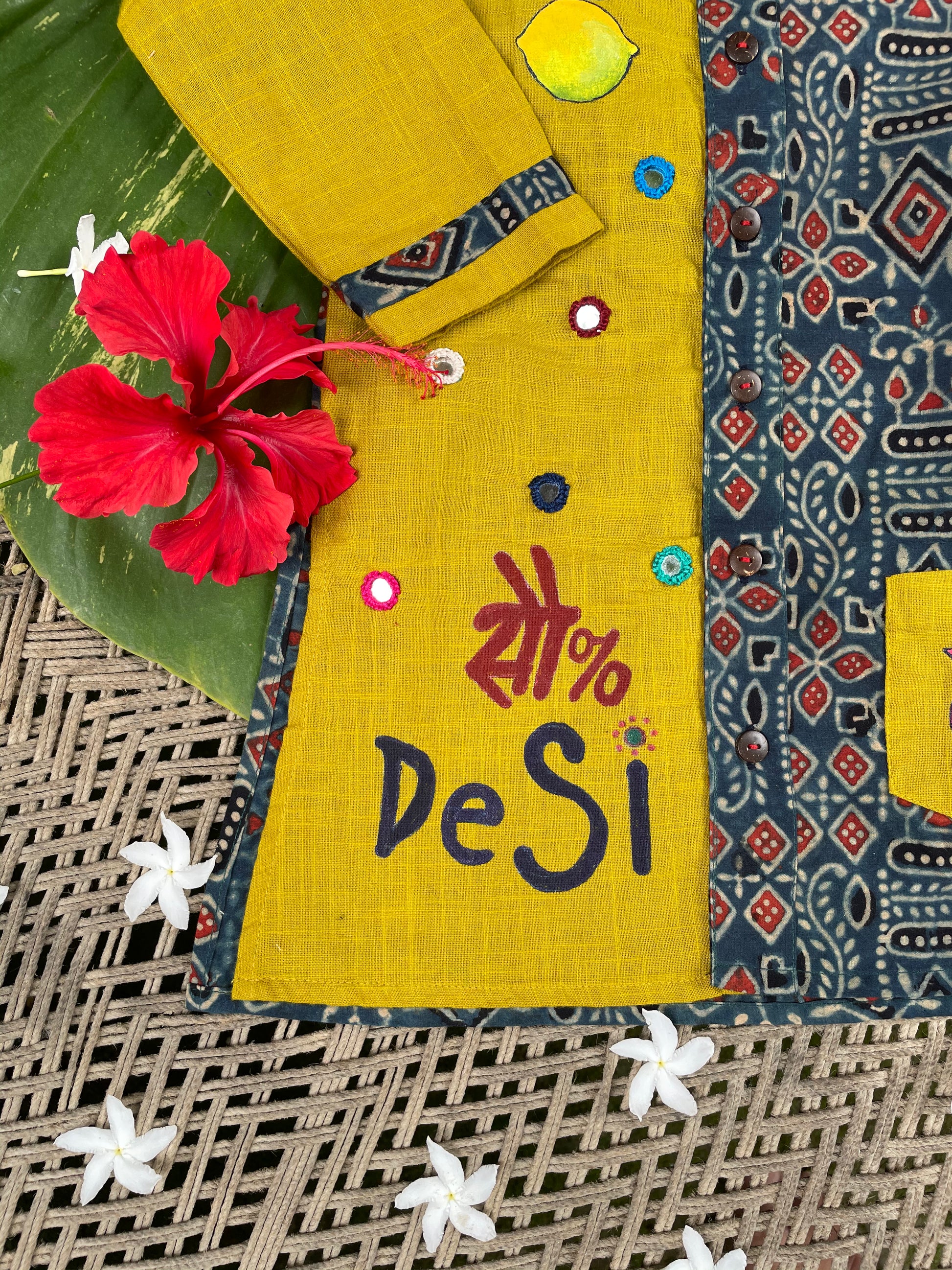 Kids traditional outfit showing a mustard yellow and indigo blue Ajrakh print kurta with handpainted mirror work, nimbu-mirchi illustration, 100% DeSi text, and white pyjamas.