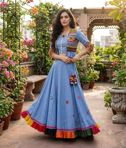 Model in a blue cotton chambray lehenga set featuring an embroidered short jacket, cowrie shell crop top, and a flared skirt with a multi-colored ruffled hem in an outdoor garden setting.