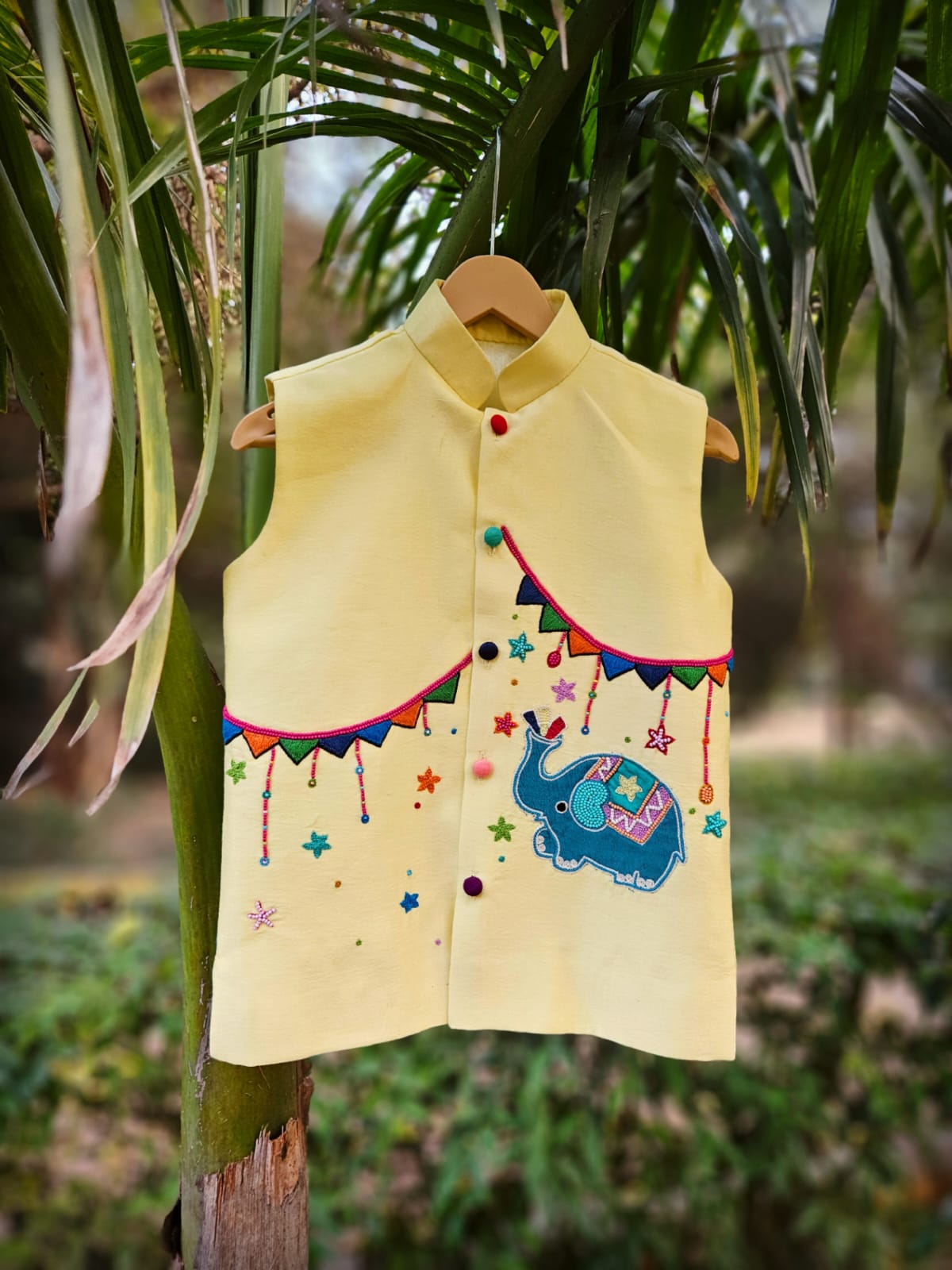 "Premium pale yellow handcrafted festive Bundi Nehru jacket for boys featuring intricate blue elephant hand embroidery and colorful pennant banner detailing."