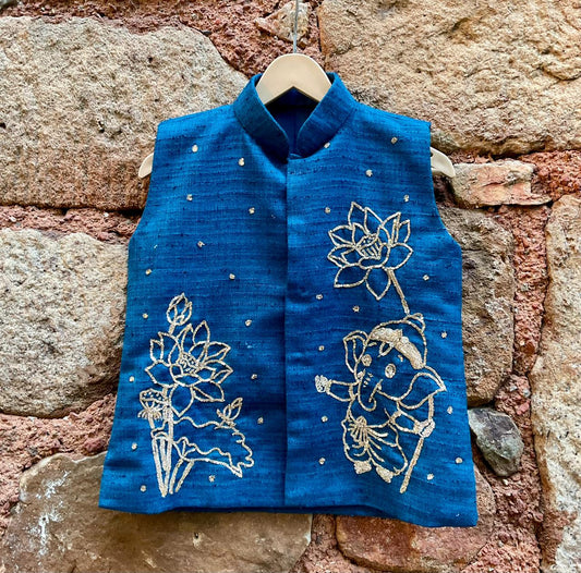Kids traditional festive wear showing a premium royal teal blue handloom cotton sleeveless Bundi jacket for boys featuring intricate gold zardosi hand embroidery of a cute baby Lord Ganesha and lotus motifs.