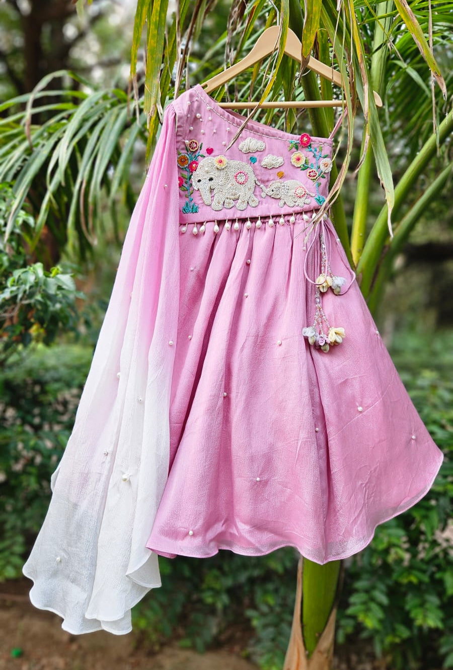 Designer pink one-shoulder girls lehenga set with embroidered elephant motif and attached ombre dupatta hanging against a green backdrop