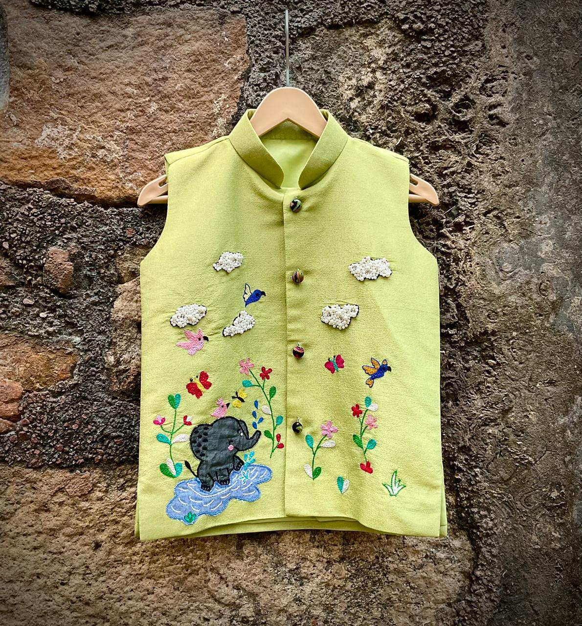 A boys' sleeveless pistachio-green ethnic Bundi jacket hanging on a wooden hanger, featuring hand-embroidered flowers, birds, butterflies, a baby elephant playing in water, and 3D beaded clouds.