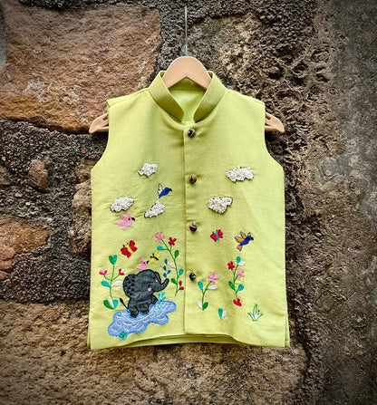 A boys' sleeveless pistachio-green ethnic Bundi jacket hanging on a wooden hanger, featuring hand-embroidered flowers, birds, butterflies, a baby elephant playing in water, and 3D beaded clouds.