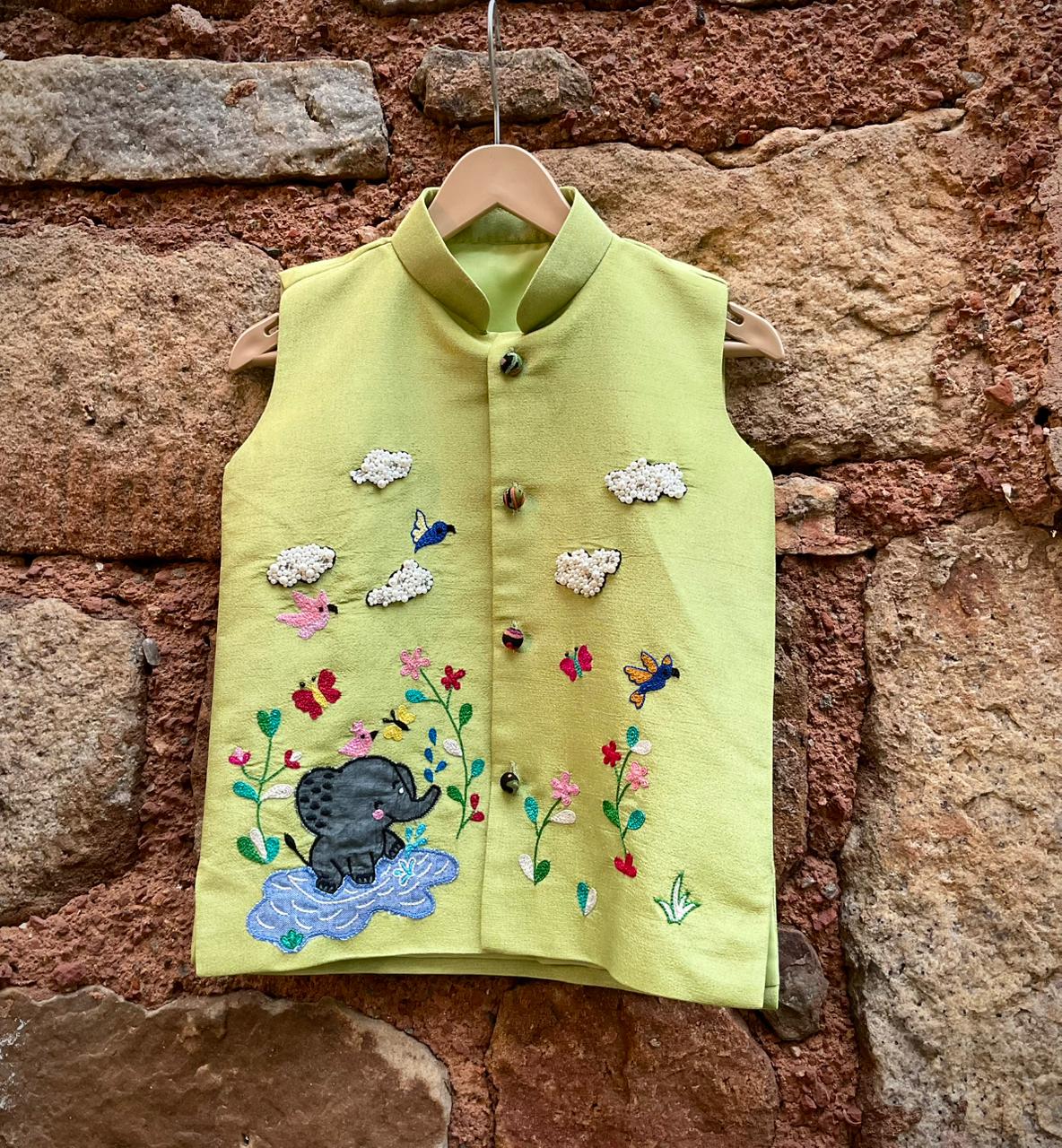 A boys' sleeveless pistachio-green ethnic Bundi jacket hanging on a wooden hanger, featuring hand-embroidered flowers, birds, butterflies, a baby elephant playing in water, and 3D beaded clouds.