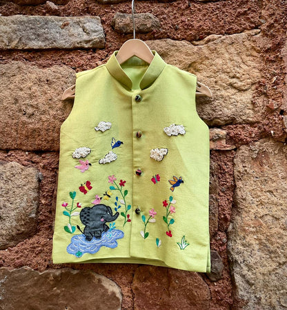 A boys' sleeveless pistachio-green ethnic Bundi jacket hanging on a wooden hanger, featuring hand-embroidered flowers, birds, butterflies, a baby elephant playing in water, and 3D beaded clouds.