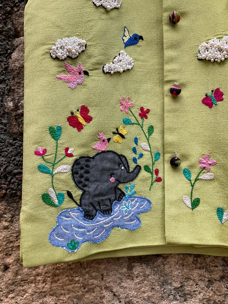 A boys' sleeveless pistachio-green ethnic Bundi jacket hanging on a wooden hanger, featuring hand-embroidered flowers, birds, butterflies, a baby elephant playing in water, and 3D beaded clouds.