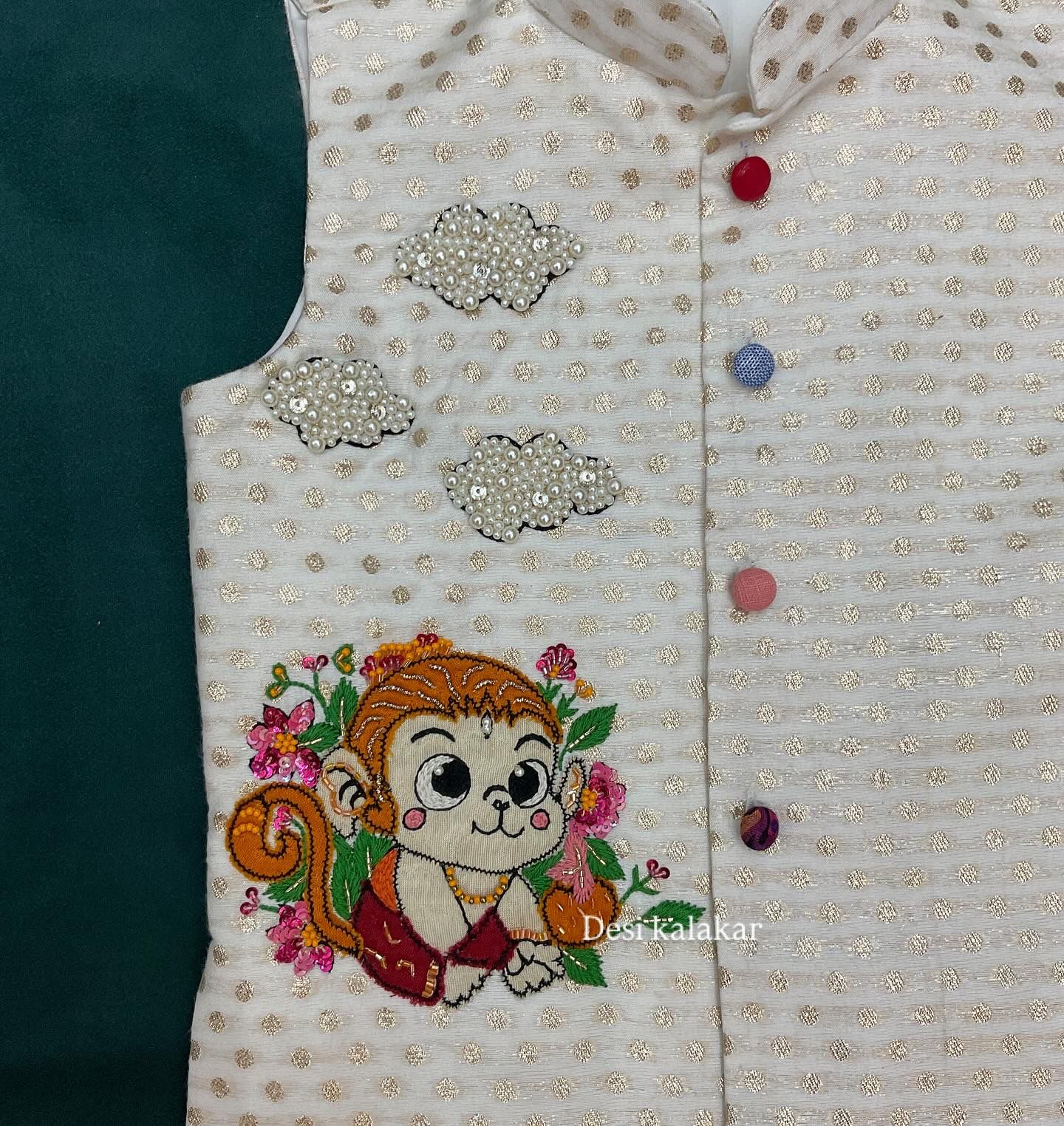 An off-white boys' ethnic Bundi jacket with woven gold zari dots, featuring a hand-embroidered cute baby Hanuman monkey character with pink flowers and three 3D pearl-beaded clouds.