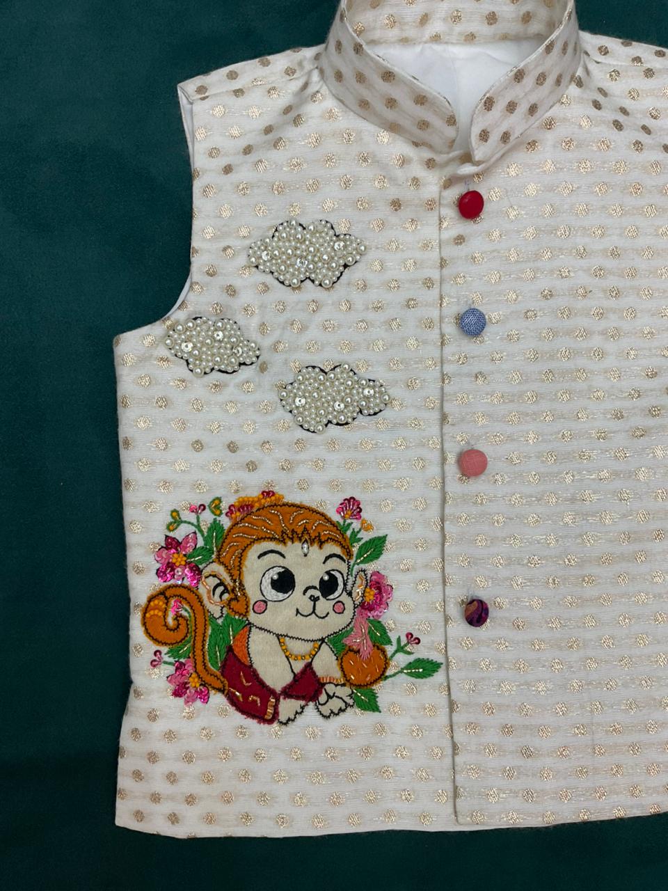 An off-white boys' ethnic Bundi jacket with woven gold zari dots, featuring a hand-embroidered cute baby Hanuman monkey character with pink flowers and three 3D pearl-beaded clouds.