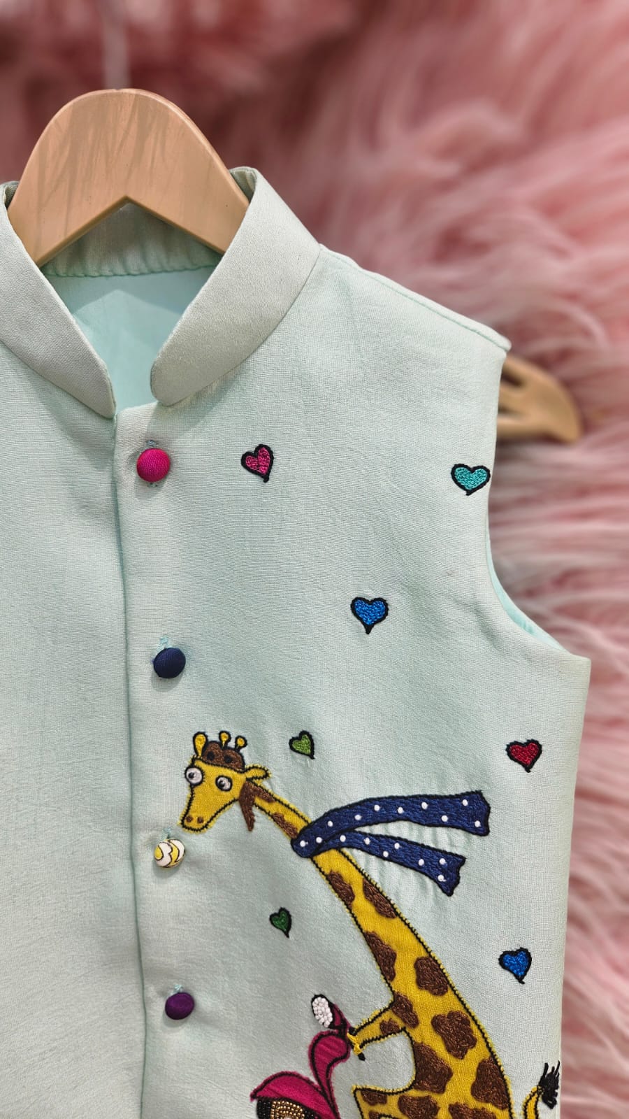 A mint green sleeveless ethnic Nehru jacket for kids hanging on a wooden hanger, featuring detailed hand-embroidery of a giraffe riding a pink scooter surrounded by tiny colorful hearts.