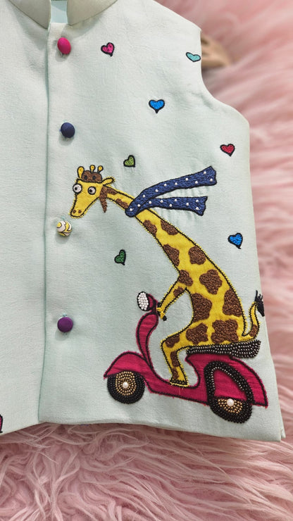 A mint green sleeveless ethnic Nehru jacket for kids hanging on a wooden hanger, featuring detailed hand-embroidery of a giraffe riding a pink scooter surrounded by tiny colorful hearts.