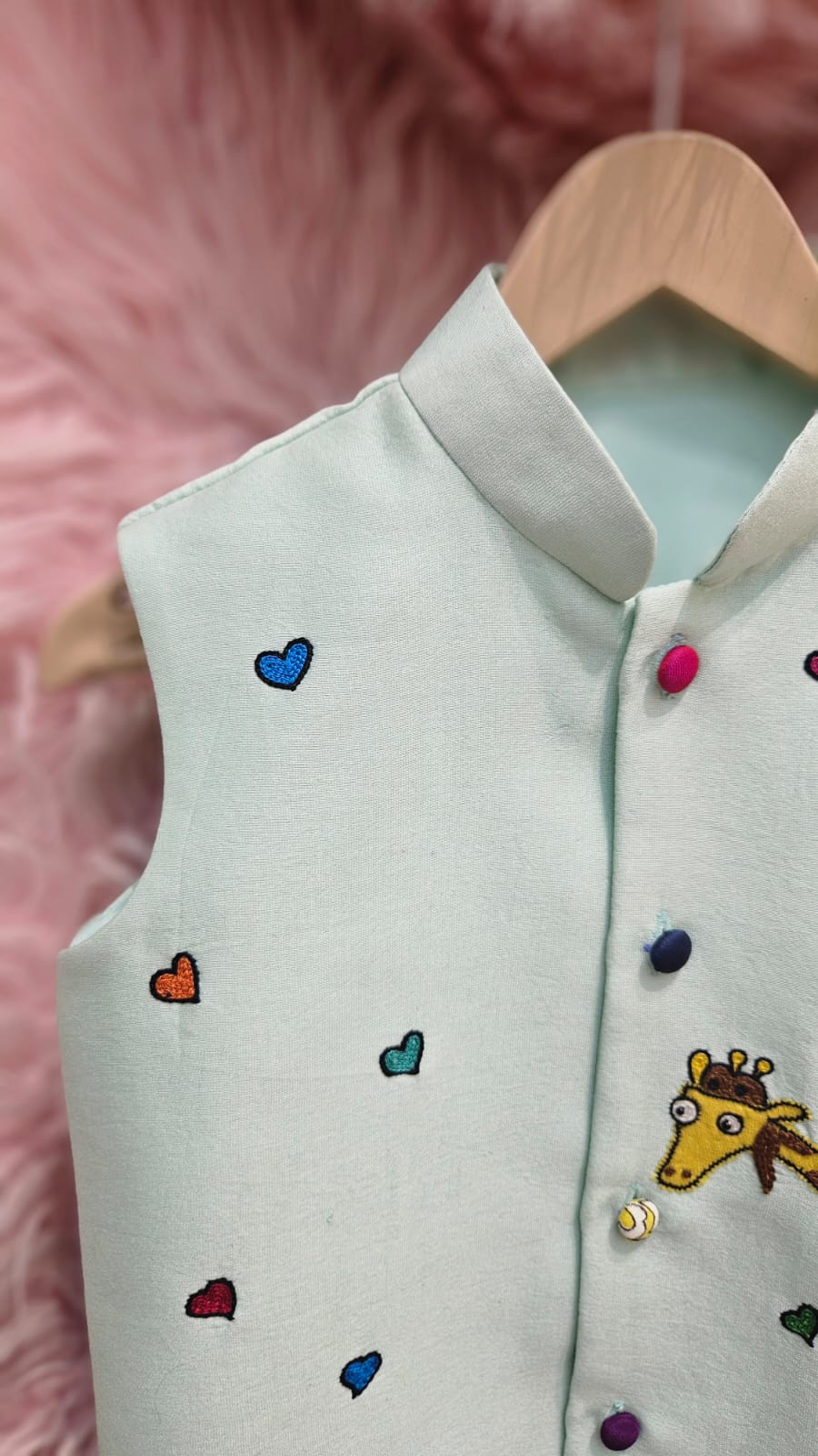 A mint green sleeveless ethnic Nehru jacket for kids hanging on a wooden hanger, featuring detailed hand-embroidery of a giraffe riding a pink scooter surrounded by tiny colorful hearts.