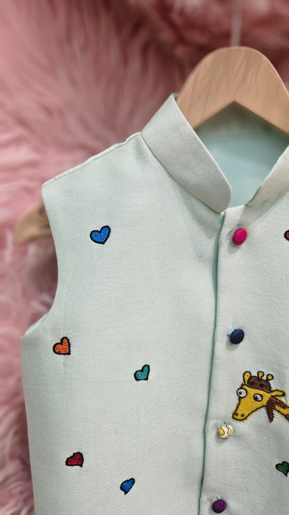 A mint green sleeveless ethnic Nehru jacket for kids hanging on a wooden hanger, featuring detailed hand-embroidery of a giraffe riding a pink scooter surrounded by tiny colorful hearts.