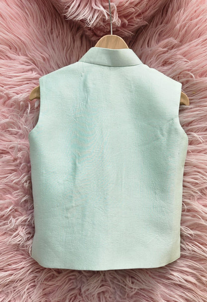 A mint green sleeveless ethnic Nehru jacket for kids hanging on a wooden hanger, featuring detailed hand-embroidery of a giraffe riding a pink scooter surrounded by tiny colorful hearts.