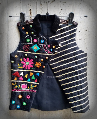 Kids traditional festive wear showing a premium black sleeveless asymmetric Bundi jacket for boys featuring multi-color Kutchi mirror work, floral embroidery, and horizontal gold stripes.