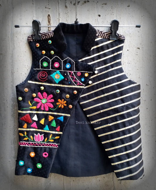 Kids traditional festive wear showing a premium black sleeveless asymmetric Bundi jacket for boys featuring multi-color Kutchi mirror work, floral embroidery, and horizontal gold stripes.