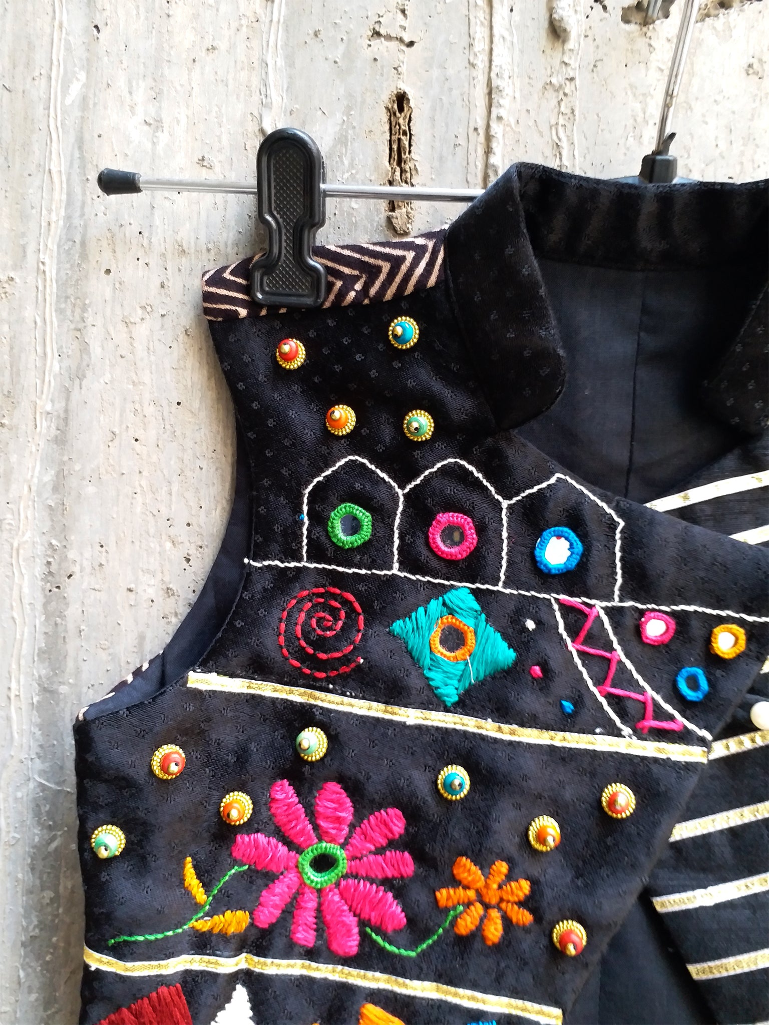 Kids traditional festive wear showing a premium black sleeveless asymmetric Bundi jacket for boys featuring multi-color Kutchi mirror work, floral embroidery, and horizontal gold stripes.