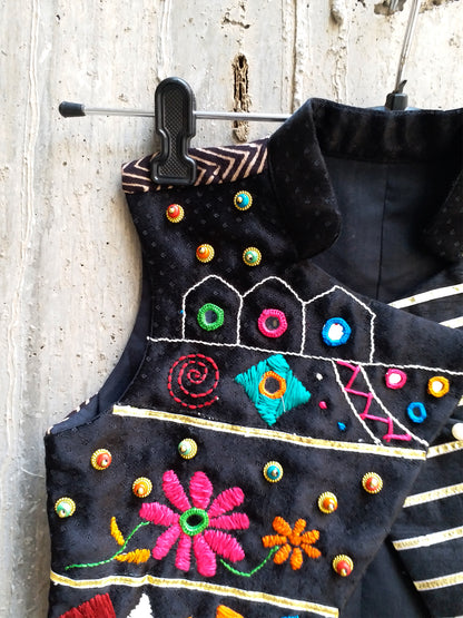 Kids traditional festive wear showing a premium black sleeveless asymmetric Bundi jacket for boys featuring multi-color Kutchi mirror work, floral embroidery, and horizontal gold stripes.