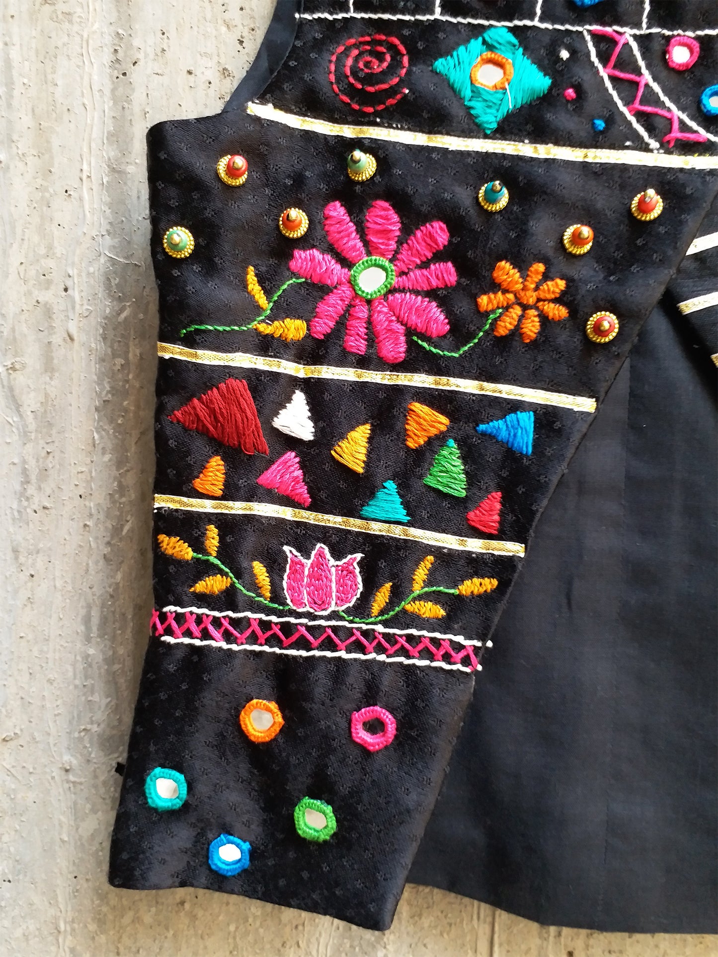 Kids traditional festive wear showing a premium black sleeveless asymmetric Bundi jacket for boys featuring multi-color Kutchi mirror work, floral embroidery, and horizontal gold stripes.