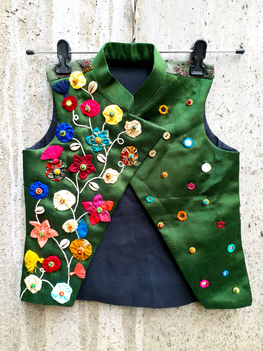Kids traditional festive wear showing an emerald green Mashru silk sleeveless asymmetric Bundi jacket for boys featuring a colorful handcrafted vine of fabric patchwork flowers, cowrie shells, tassels, and scattered multi-color mirror work details.