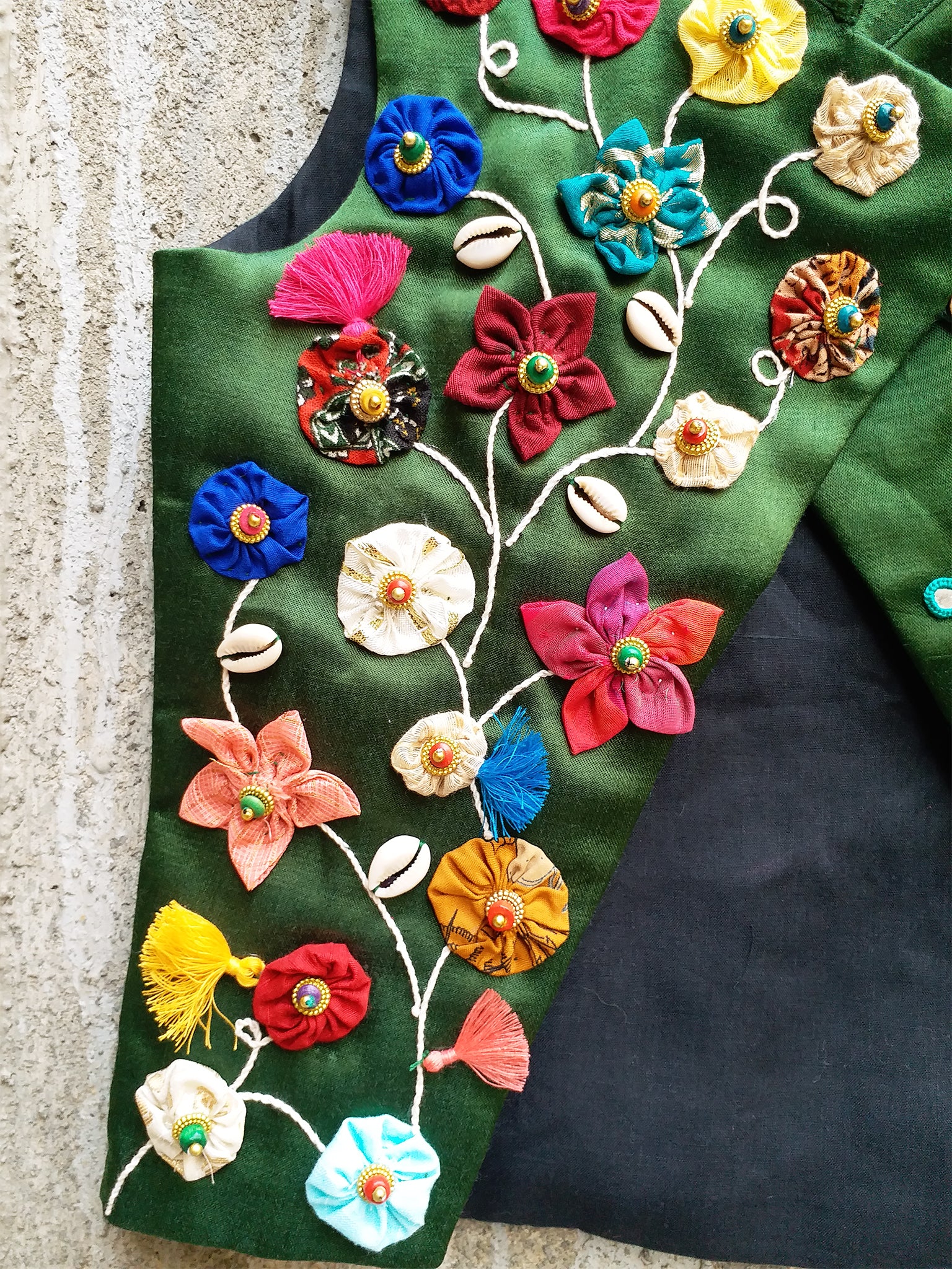 Kids traditional festive wear showing an emerald green Mashru silk sleeveless asymmetric Bundi jacket for boys featuring a colorful handcrafted vine of fabric patchwork flowers, cowrie shells, tassels, and scattered multi-color mirror work details.